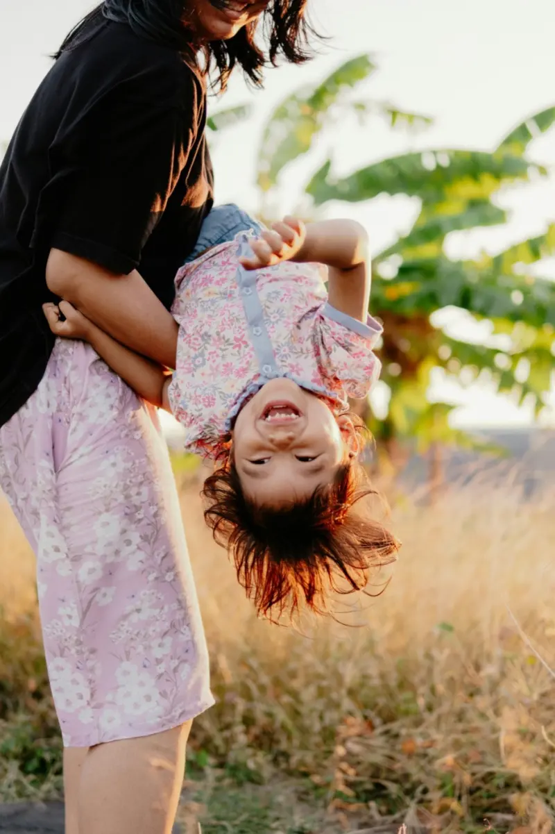 Une maman qui fait rire sa fille la tête en bas, dans un champ au soleil